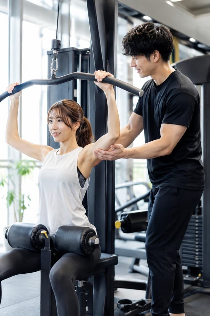 男性トレーナーが女性のラットプルダウンのフォームをサポートしている様子